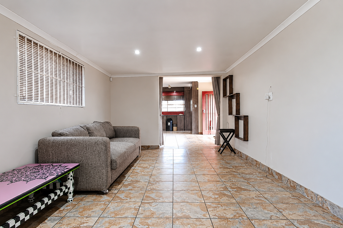 A spacious living area featuring tiled flooring, recessed ceiling lighting, and a window with fitted blinds, leading through to an adjacent kitchen space.
