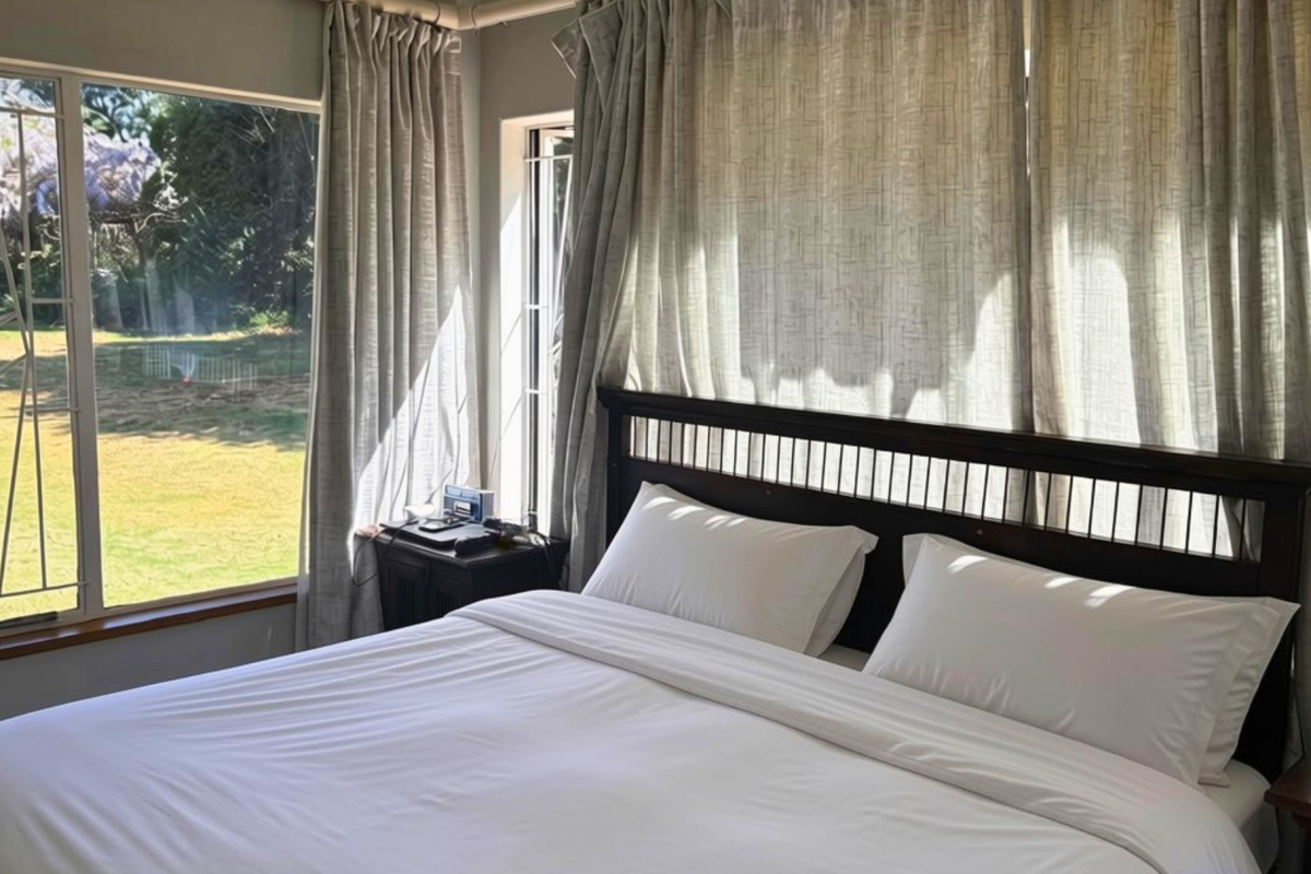 A bright bedroom featuring large windows with security bars, offering natural light and views of the garden.