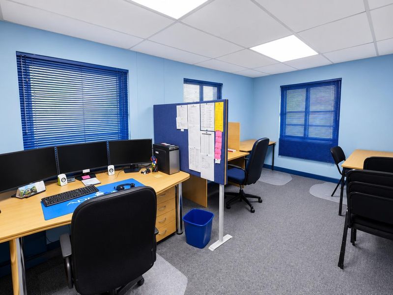 A functional office space featuring carpeted flooring, suspended ceiling with recessed lighting, and blue walls with fitted venetian blinds.