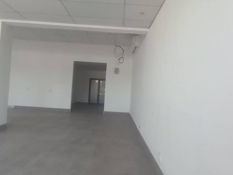 An empty, open-plan retail space featuring tiled flooring, a suspended ceiling with recessed lighting, and a structural column.