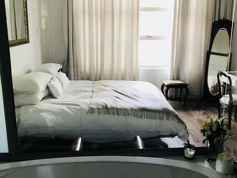 A well-lit bedroom featuring laminate flooring and a large window, with a view into an adjacent bathroom area containing a built-in bathtub.