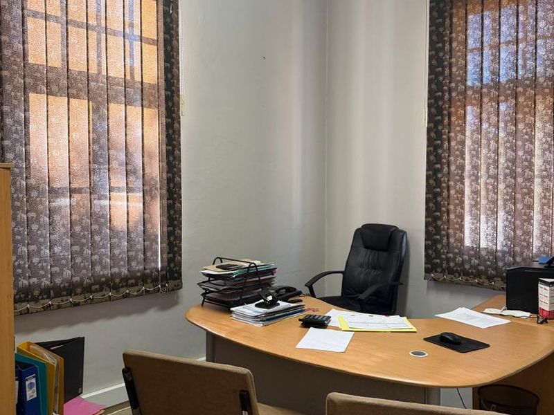 A functional home office space featuring carpeted flooring, neutral wall finishes, and dual windows fitted with vertical blinds.