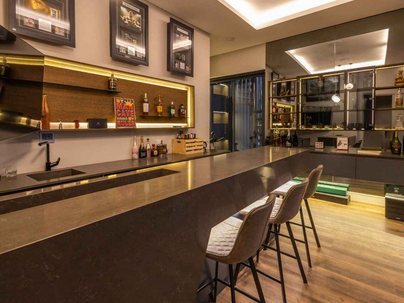 A modern, high-end home bar featuring a sleek stone countertop, integrated sink, ambient LED lighting, and sophisticated mirrored wall accents.