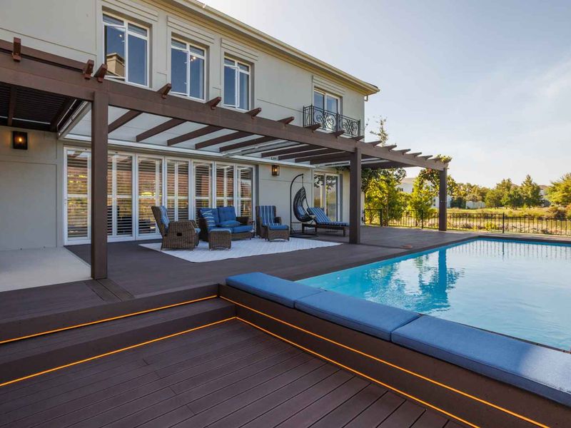 A well-maintained outdoor entertainment area featuring a swimming pool, timber decking with integrated lighting, and a pergola, connected to the home via folding doors.