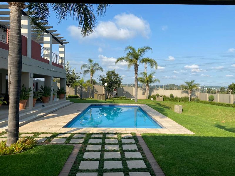 A well-maintained outdoor pool area featuring a swimming pool, paved deck, covered patio with pergola, and landscaped garden surroundings.