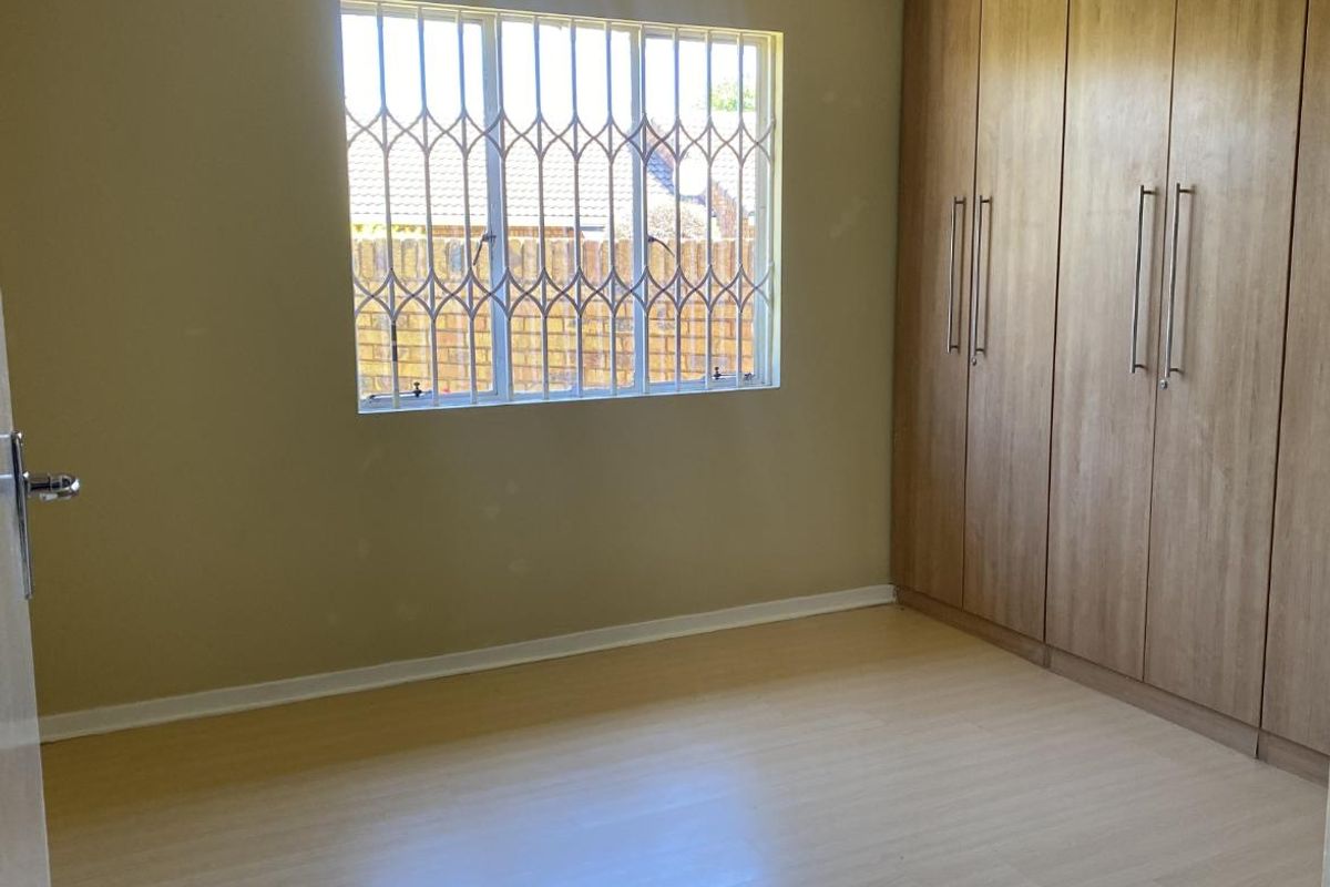 A clean, empty bedroom featuring light-toned laminate flooring and floor-to-ceiling built-in wooden cupboards, with security bars installed on the window.