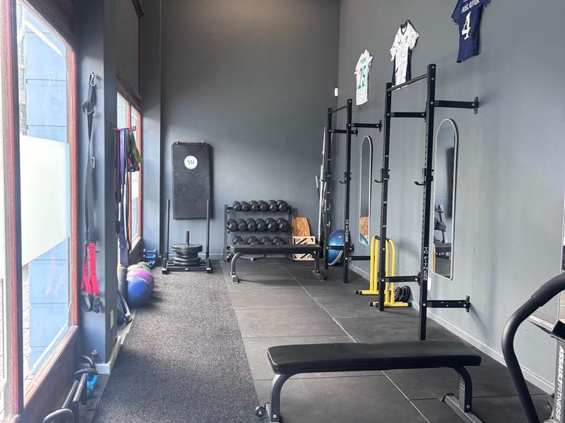 A functional gym space featuring rubberized flooring, wall-mounted squat racks, and large windows providing natural light.