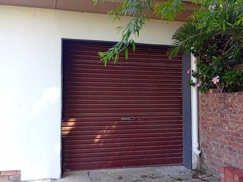 A single garage featuring a red roller shutter door and a concrete driveway, situated next to a brick retaining wall.