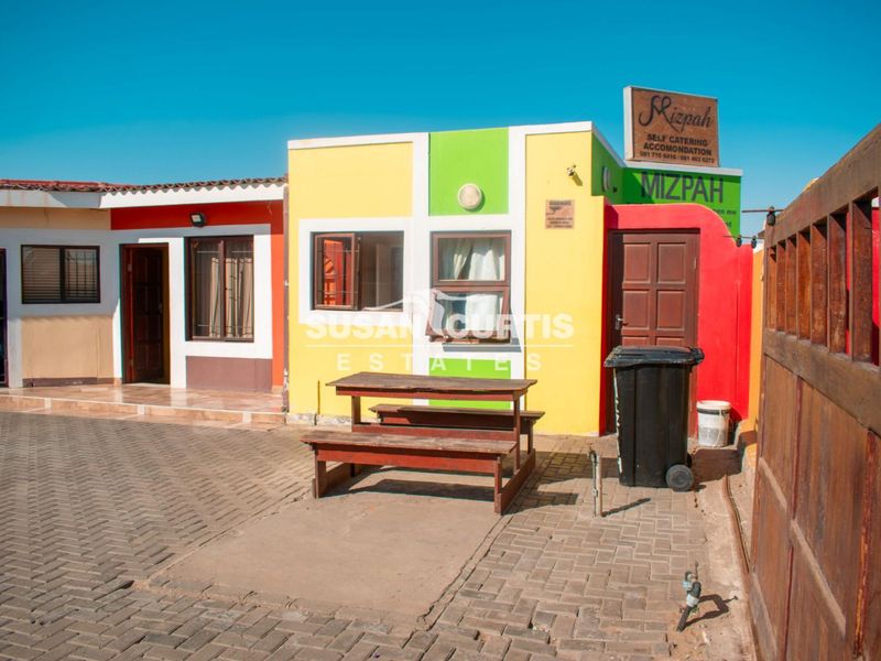 Front exterior view of the guest house featuring a paved driveway, timber gate, and brightly painted facade with signage.