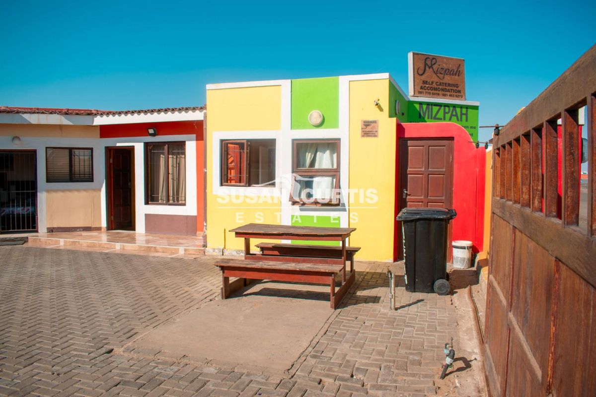 Front exterior view of the guest house featuring a paved driveway, timber gate, and brightly painted facade with signage.
