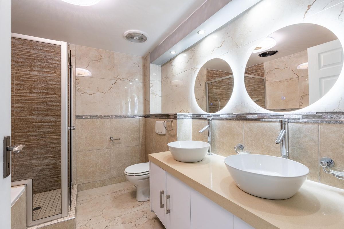 Modern bathroom featuring a double vanity with vessel sinks, backlit circular mirrors, and a spacious walk-in shower with neutral marble-style tiling.