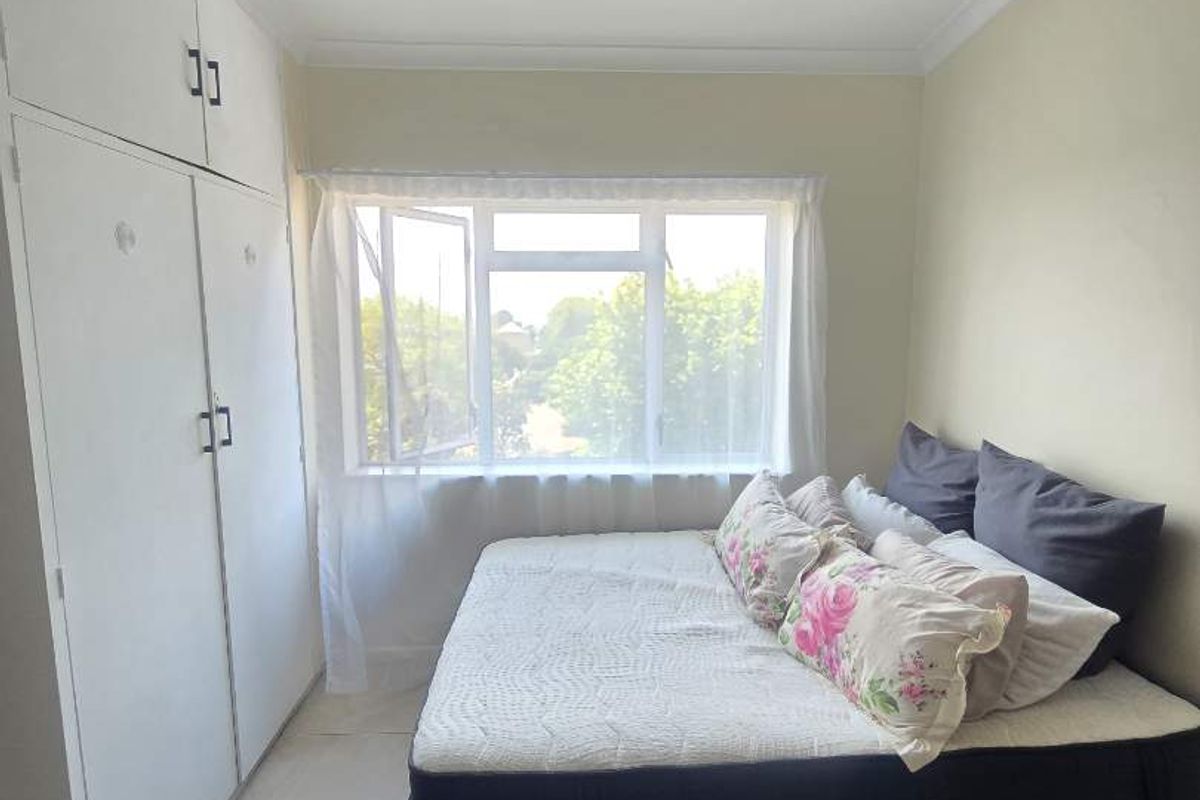 A functional bedroom featuring built-in cupboards, neutral tiled flooring, and a large window providing natural light.