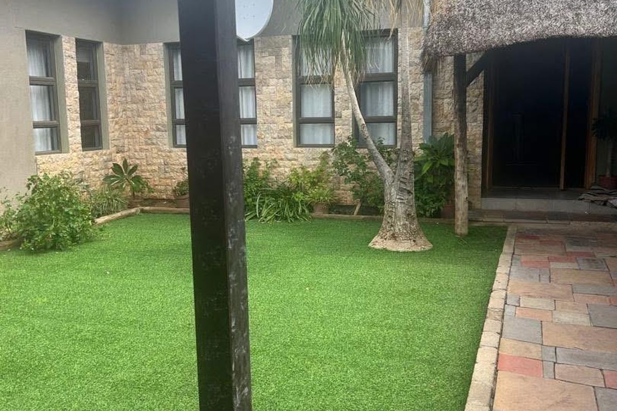 A well-maintained rear courtyard featuring a paved walkway, artificial lawn, stone-clad exterior walls, and a thatched roof entrance.