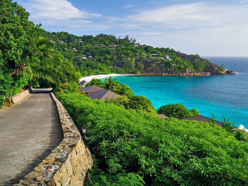 A scenic view of a coastal road winding along a lush hillside overlooking a turquoise bay and sandy beach.