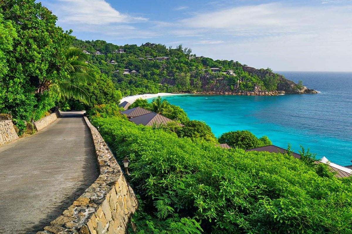 A scenic view of a coastal road winding along a lush hillside overlooking a turquoise bay and sandy beach.