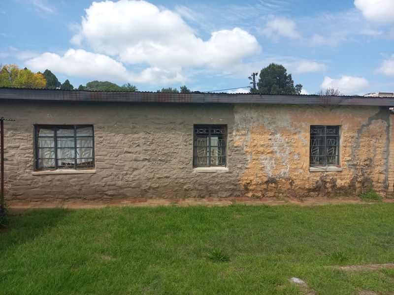 Rear exterior view of a stone masonry house featuring metal security window grilles and a corrugated metal roof showing signs of weathering.