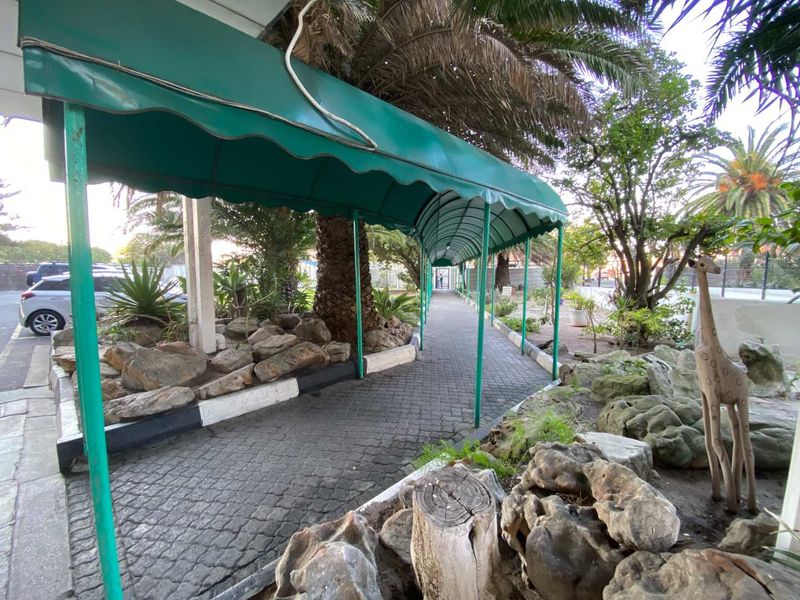 A covered paved walkway leads through a landscaped garden area with mature trees and decorative rock features at the property entrance.