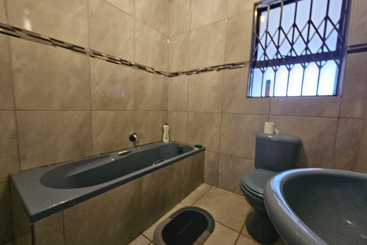 A functional bathroom featuring a built-in bathtub, toilet, and washbasin with neutral wall and floor tiling and a security-fitted window.