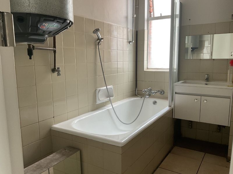 A dated bathroom featuring a bathtub with shower mixer, wall-mounted geyser, and vanity basin, requiring modernization of finishes and fixtures.