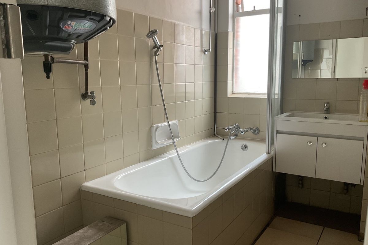 A dated bathroom featuring a bathtub with shower mixer, wall-mounted geyser, and vanity basin, requiring modernization of finishes and fixtures.