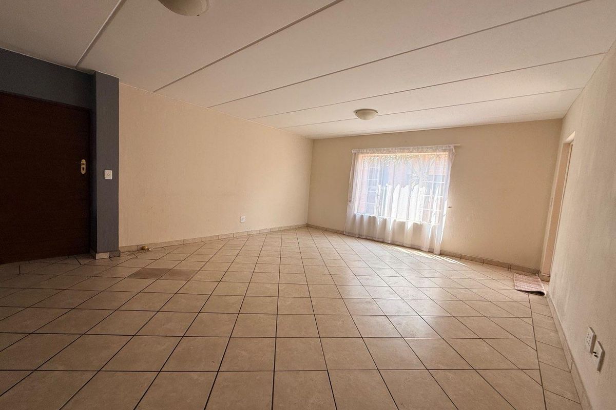 A spacious, empty living area featuring neutral tiled flooring, a large window with sheer curtains, and a simple ceiling-mounted light fixture.