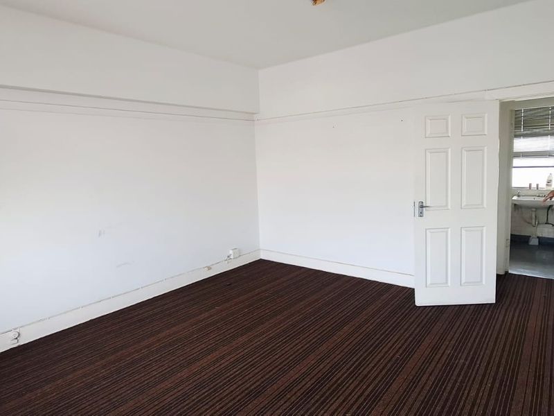 An empty bedroom featuring dark striped carpeting and white walls, with an adjacent small wash area visible through the open doorway.