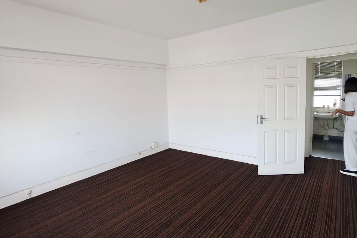 An empty bedroom featuring dark striped carpeting and white walls, with an adjacent small wash area visible through the open doorway.