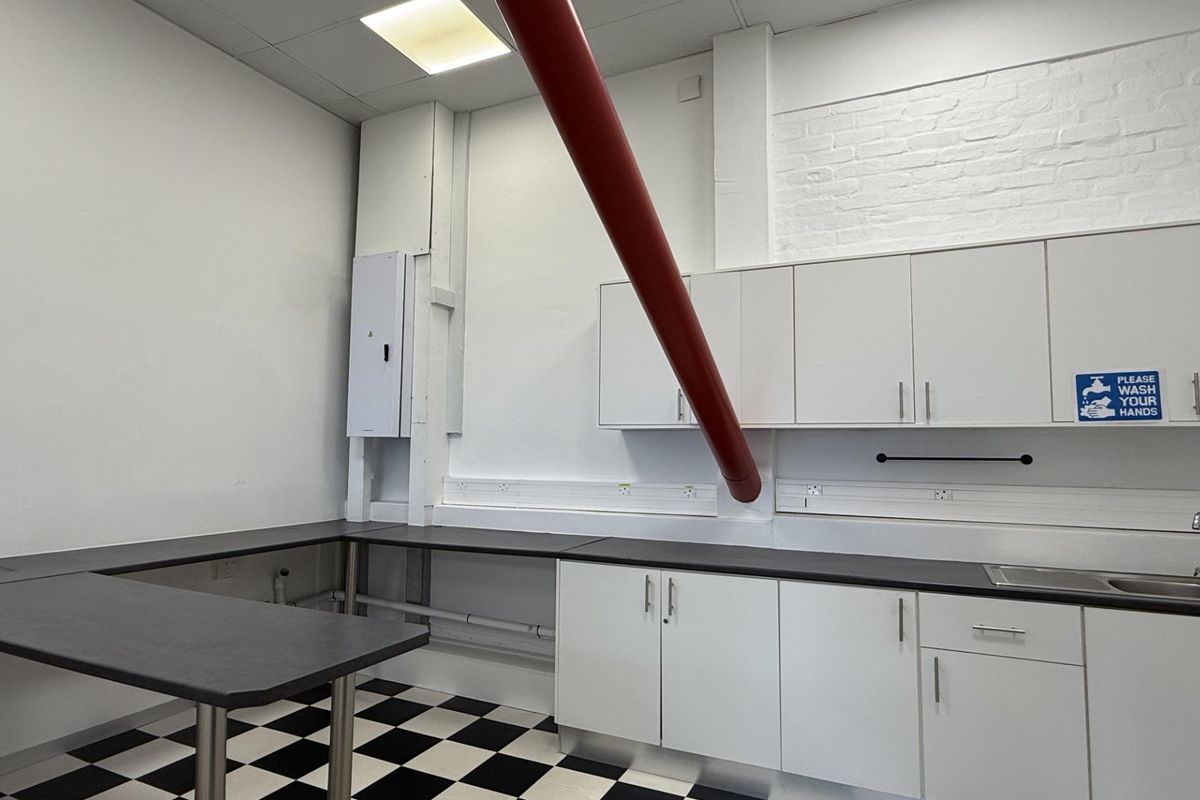 A functional office kitchenette featuring white cabinetry, dark laminate countertops, a stainless steel sink, and a distinctive checkerboard floor pattern.