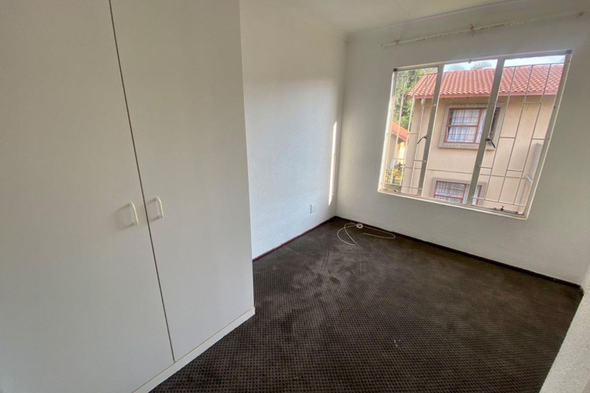 A vacant bedroom featuring built-in cupboards, dark carpeting, and a window with security bars overlooking the complex.