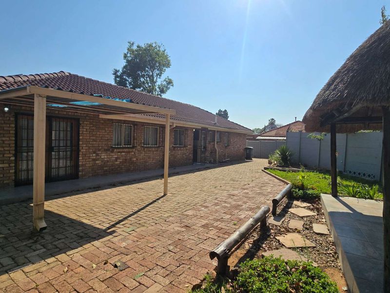 Rear exterior view featuring a brick house, paved patio area, and a traditional thatched lapa structure in the garden.
