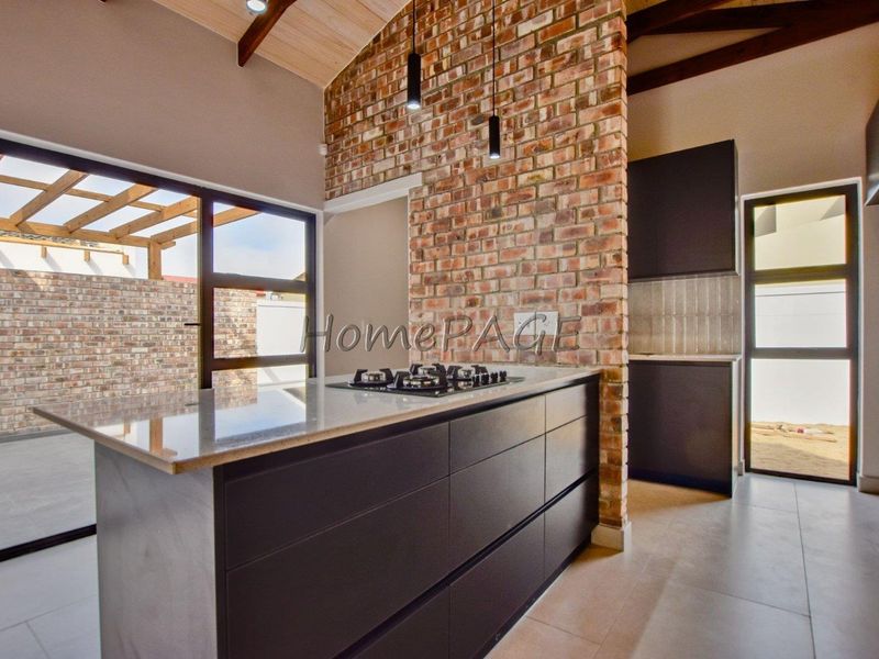 Modern kitchen featuring an exposed brick wall, central island with gas hob, stone countertops, and vaulted timber ceilings with sleek pendant lighting.