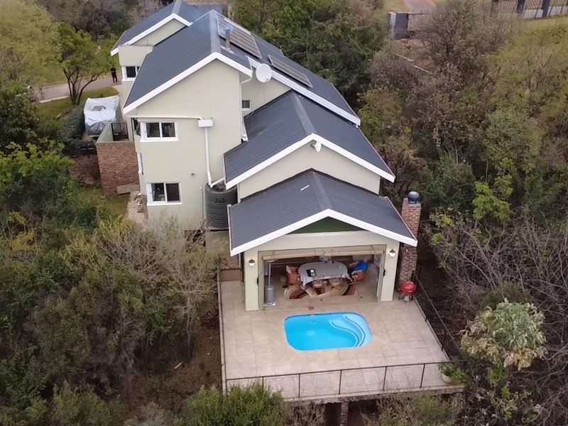 Aerial view of a multi-story house featuring a metal roof, solar panels, a covered patio, and a swimming pool set within a wooded area.