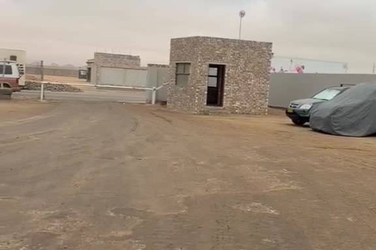 Industrial property entrance featuring a stone-clad security guard house, perimeter walling, and an access gate on an unpaved site.