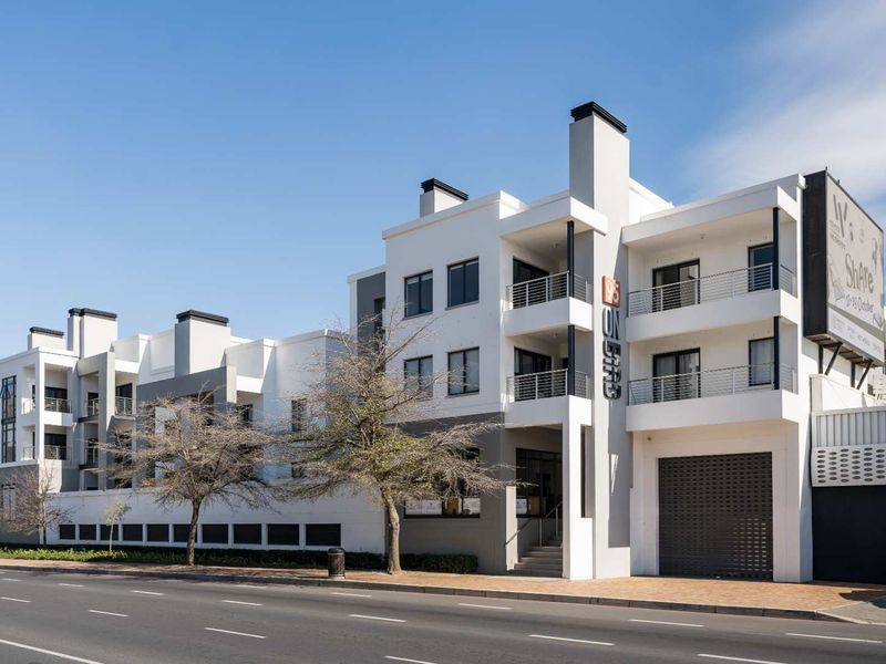 Modern multi-story apartment building facade featuring white and grey exterior finishes, private balconies with metal railings, and a ground-level roller shutter garage entrance.