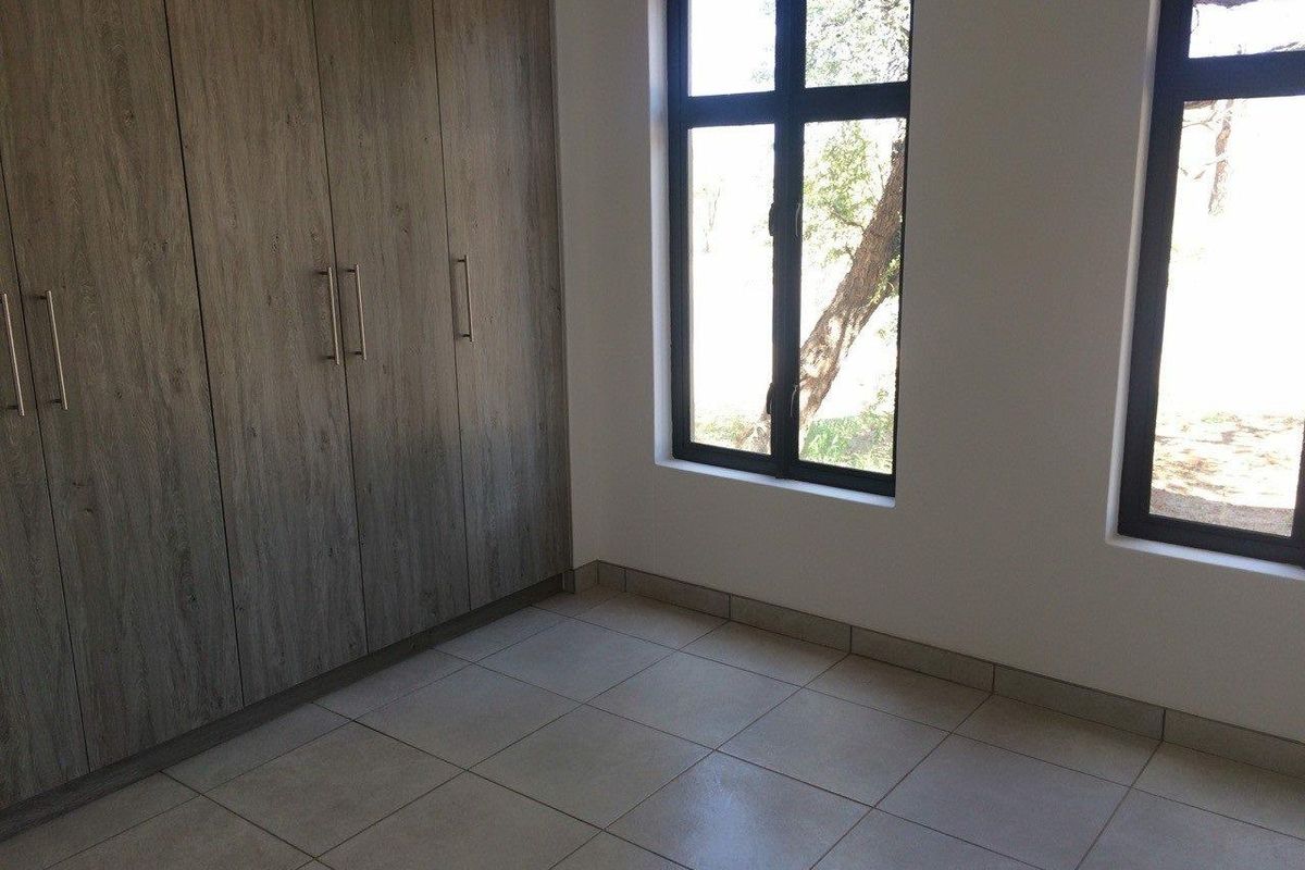 A bright, empty bedroom featuring modern wood-finish built-in cupboards, tiled flooring, and large windows with dark frames.