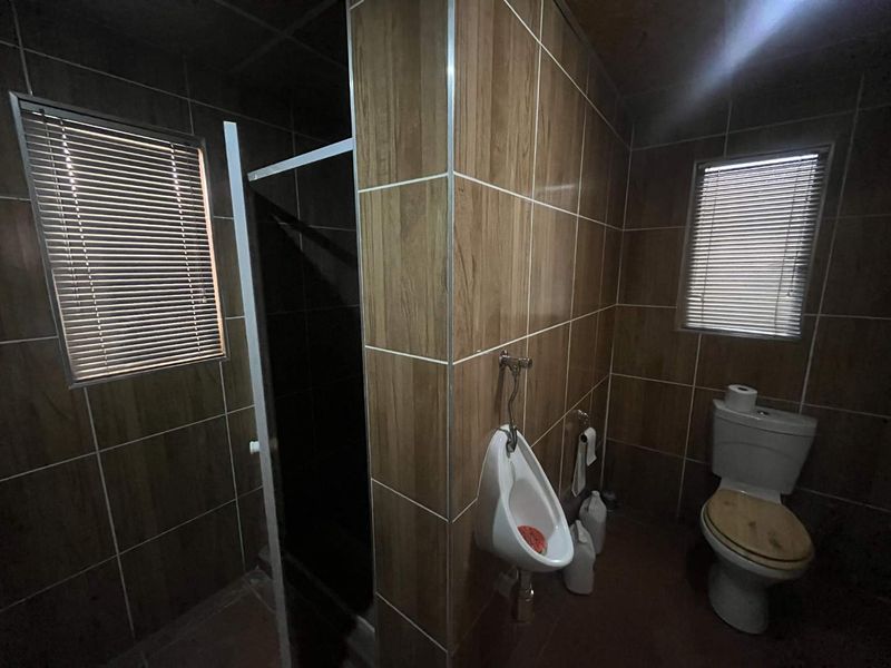 A functional bathroom featuring a urinal, toilet, and a separate shower enclosure with wood-look wall tiling and venetian blinds.