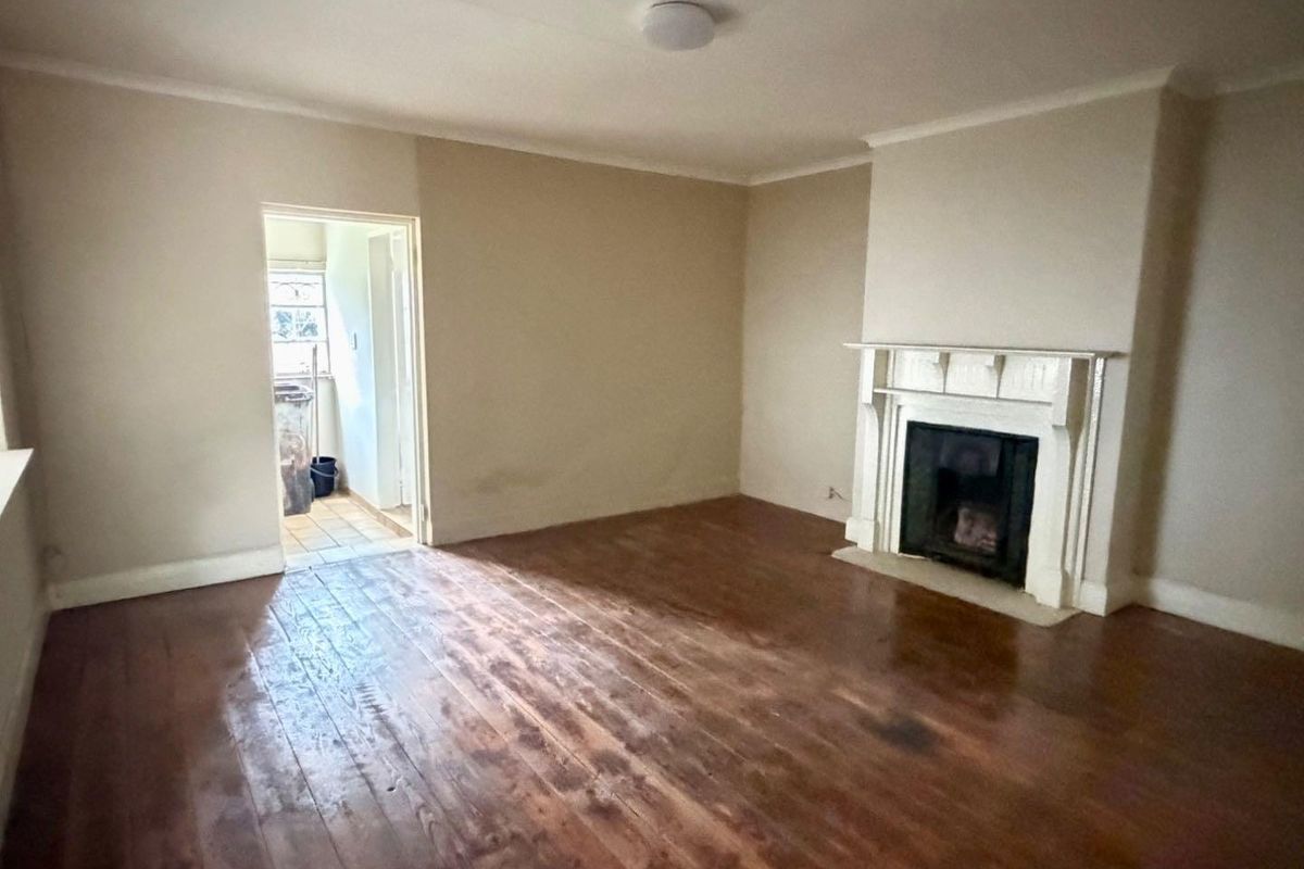 An empty living room featuring original timber flooring and a traditional fireplace mantel, with an adjacent doorway leading to a utility area.