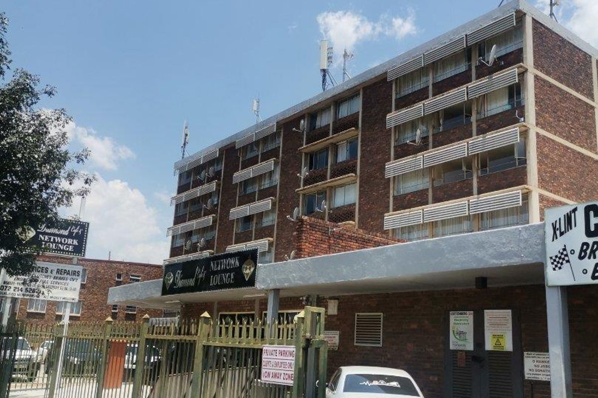 Exterior view of a multi-story brick apartment building featuring a gated entrance, security fencing, and various commercial signage on the premises.