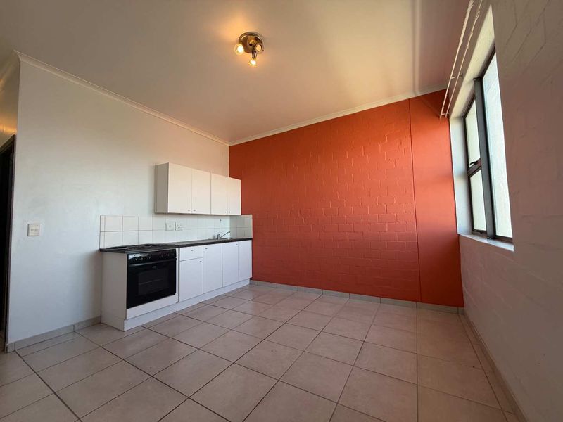A compact, empty kitchen featuring white built-in cabinetry, a built-in oven and hob, tiled flooring, and a distinctive orange brick feature wall.