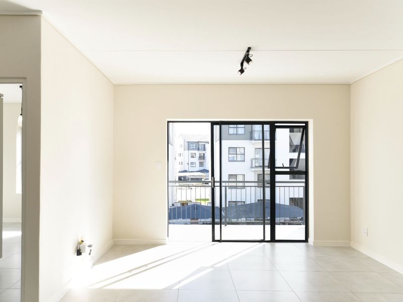 A bright, empty living area featuring neutral tiled floors, modern track lighting, and a large sliding glass door leading to a balcony.