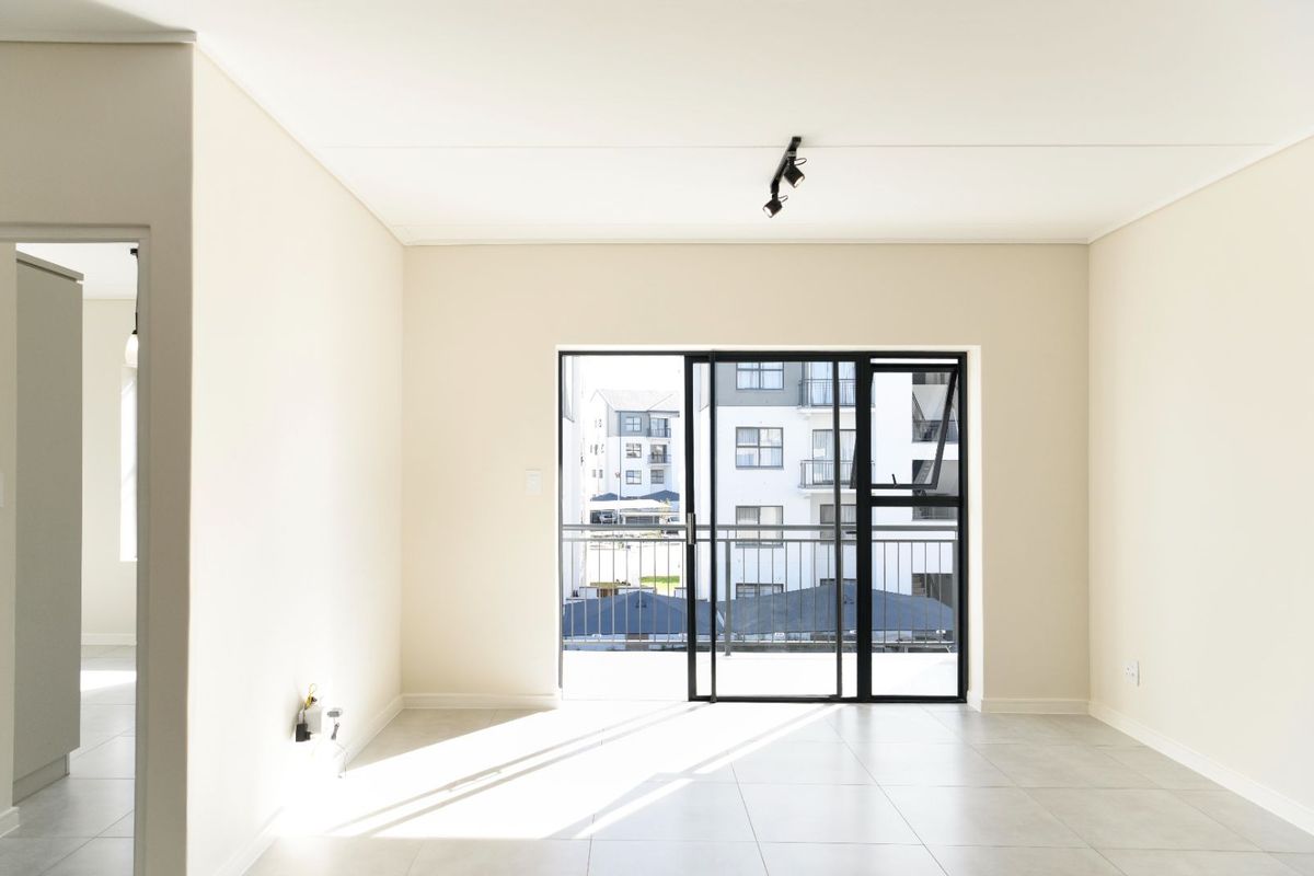 A bright, empty living area featuring neutral tiled floors, modern track lighting, and a large sliding glass door leading to a balcony.