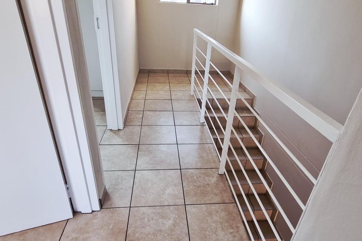 A bright upper-level hallway featuring tiled flooring and a white metal staircase railing, providing access to adjacent rooms.
