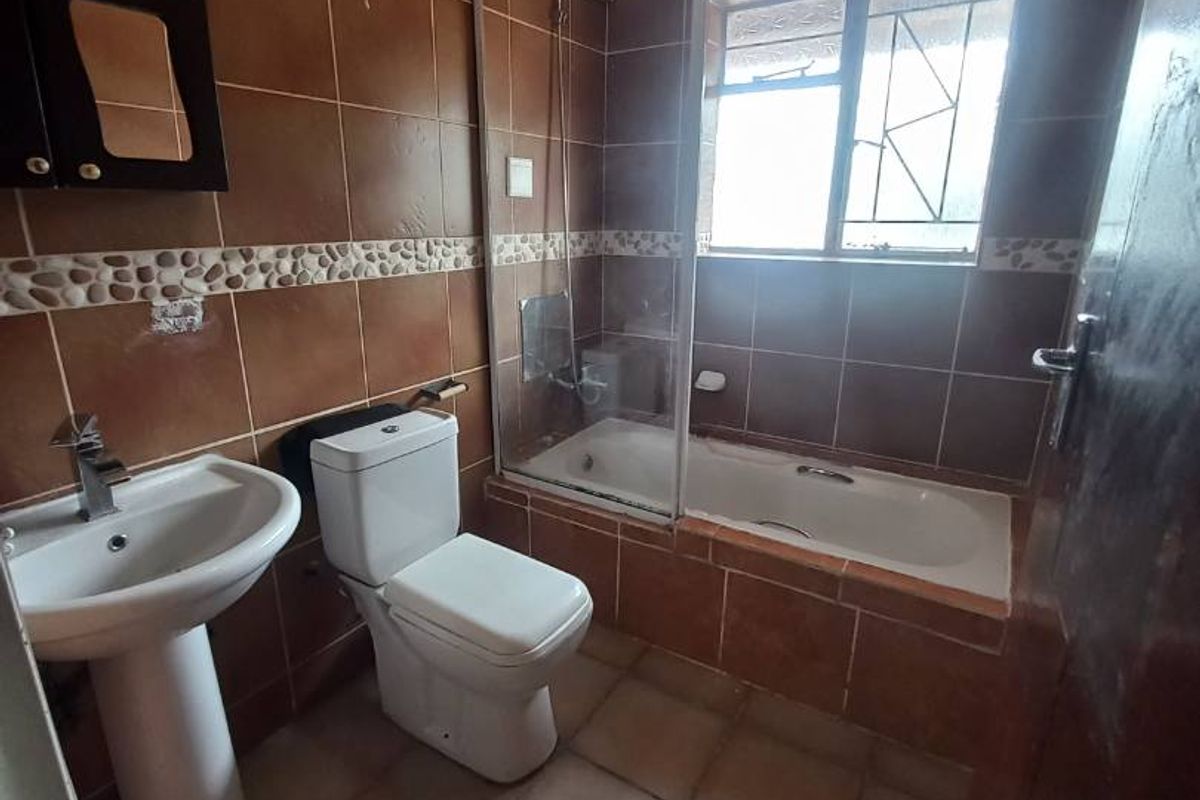 A functional bathroom featuring a pedestal basin, toilet, and a bathtub with a glass shower screen, finished with full-height brown wall tiling.
