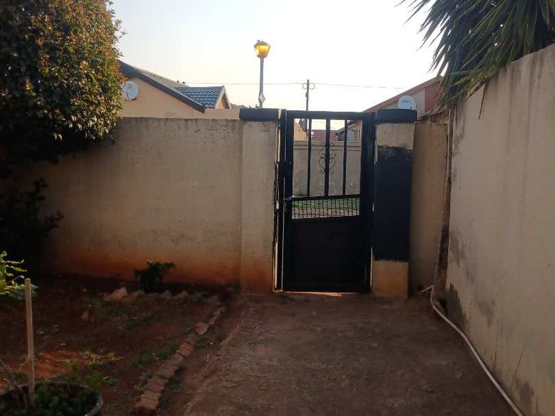A rear outdoor area featuring a concrete paved path, perimeter wall, and a metal pedestrian gate leading to the exterior.
