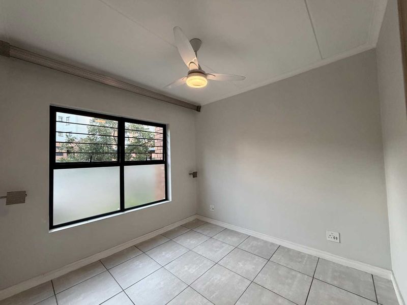 A clean, empty bedroom featuring neutral tiled flooring, a modern ceiling fan, and an aluminium-framed window with security bars.