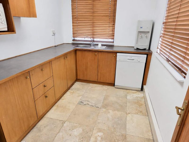 A functional office kitchenette featuring wood-finish cabinetry, laminate countertops, a stainless steel sink, and tiled flooring with natural light from dual windows.