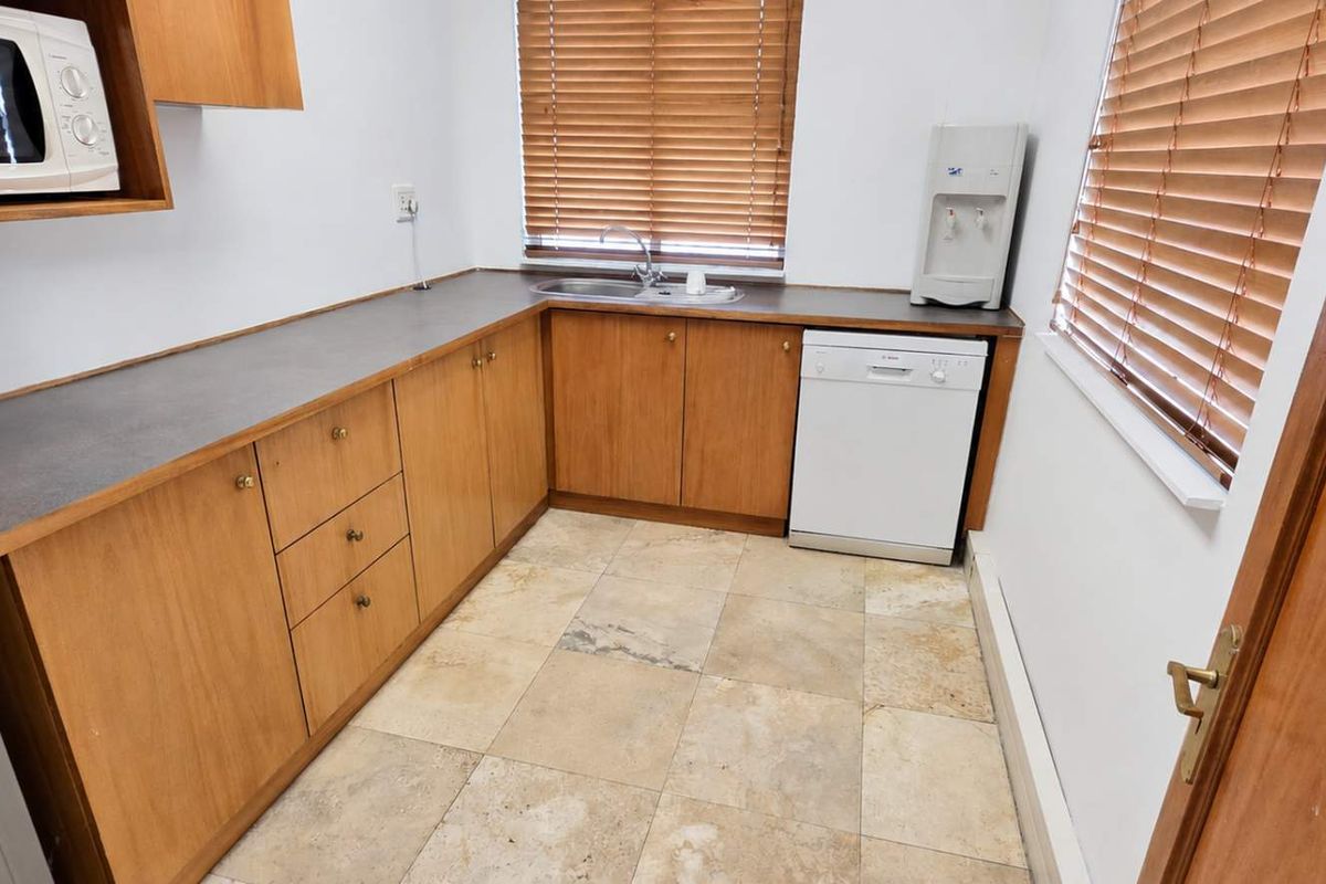 A functional office kitchenette featuring wood-finish cabinetry, laminate countertops, a stainless steel sink, and tiled flooring with natural light from dual windows.