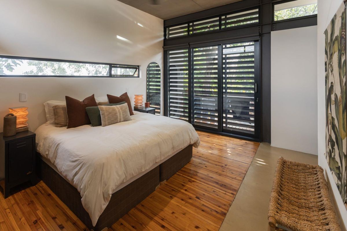 Modern bedroom featuring timber flooring, exposed steel structural elements, and large sliding glass doors with integrated louvre shutters providing natural light and outdoor access.