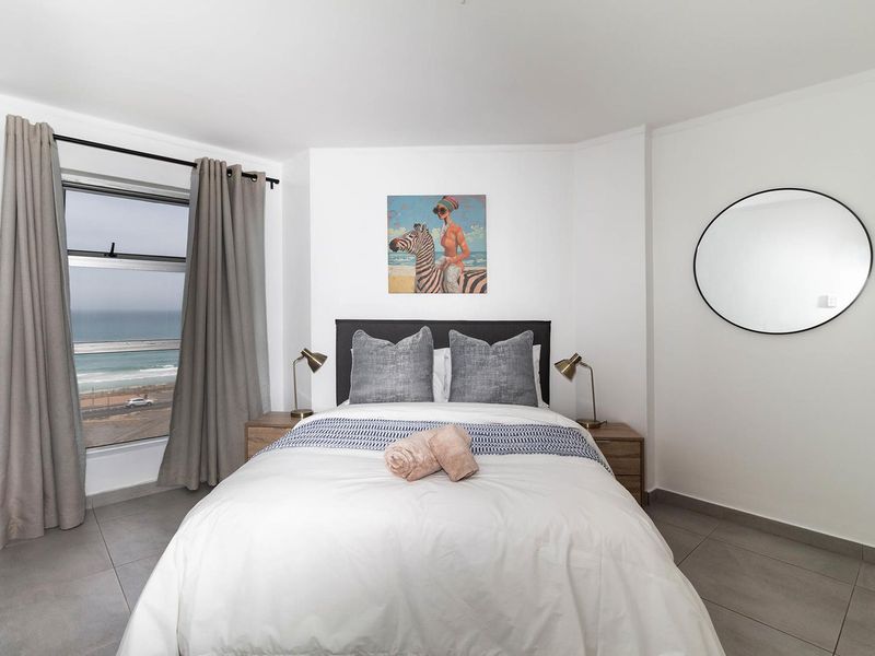 A bright bedroom featuring neutral tiled flooring and a large window offering scenic coastal views.