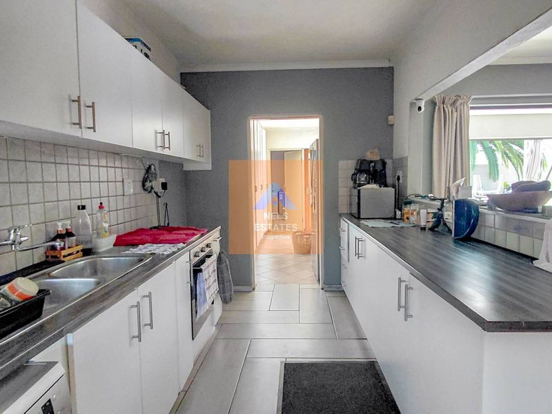 A functional galley-style kitchen featuring white cabinetry, dark laminate countertops, a double stainless steel sink, and tiled flooring.
